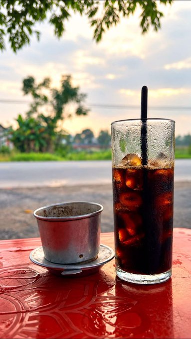 A glass of iced coffee with a straw sits on a red outdoor table alongside a metal cup, with a sunset sky and green trees in the background.