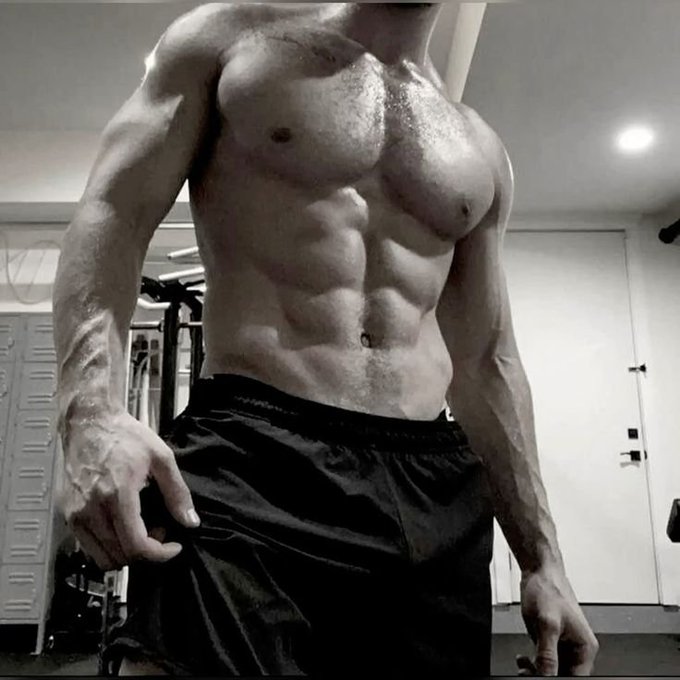 A shirtless man with a highly defined, muscular physique taking a gym mirror selfie in black shorts.