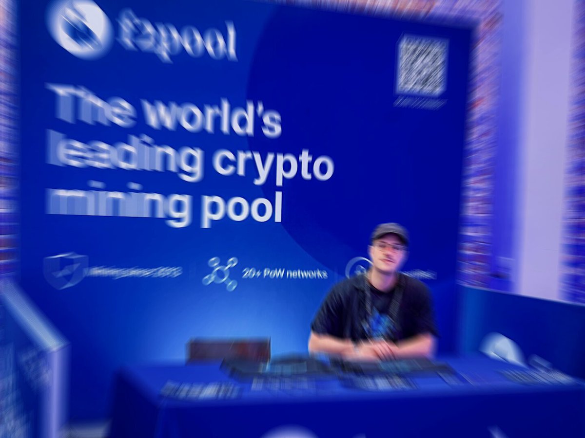 The image features a man sitting behind a desk, possibly at an event or convention related to cryptocurrency. He appears to be wearing a hat and has a camera around his neck. The background of the image shows a blue banner with white text that reads "The world's leading crypto mining pool". This suggests that the man might be involved in promoting or discussing cryptocurrency-related activities, such as mining pools for Bitcoin transactions.