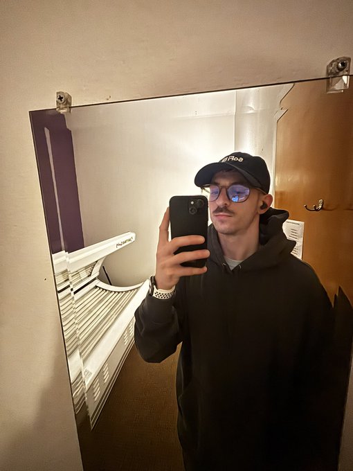 A young man wearing glasses, a mustache, a black hoodie, and a cap takes a mirror selfie with a smartphone in what appears to be a bathroom.