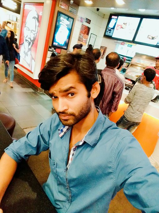 A selfie of a young man with dark hair and a beard wearing a blue shirt, taken inside what appears to be a KFC restaurant with menu boards visible in the background.