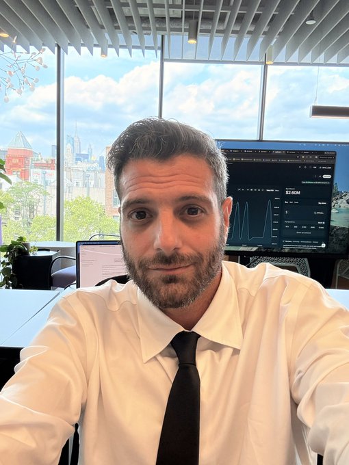 A selfie of a bearded man in a white shirt and black tie sitting at a desk in a modern office with a large window, with a computer monitor behind him displaying analytics with the figure '82.40M'.