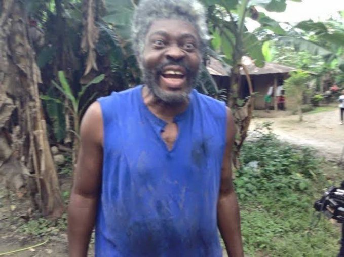 The image features a man wearing a blue shirt, standing outdoors with his mouth open wide as if he's laughing or making a funny face for the camera. The surrounding environment includes trees and bushes, giving the scene an outdoor setting. There are also some people in the background who appear to be engaged in conversation or other activities.