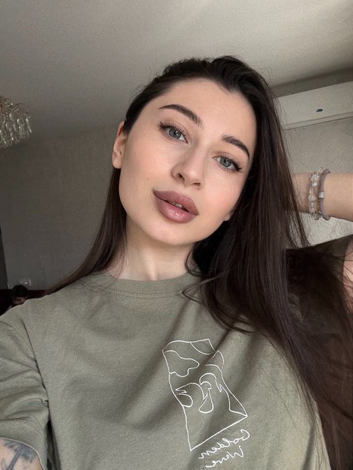 A close-up selfie of a young woman with long dark hair wearing an olive green t-shirt with a graphic design, posing with her hand behind her head showing beaded bracelets.