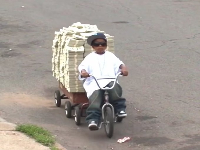 A young boy wearing sunglasses pedals a small tricycle down a street while pulling a red wagon loaded with a massive stack of cash bound by rubber bands.
