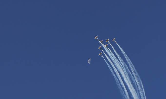 A formation of jets flying through a clear blue sky leaving white smoke trails, with a crescent moon visible in the background.