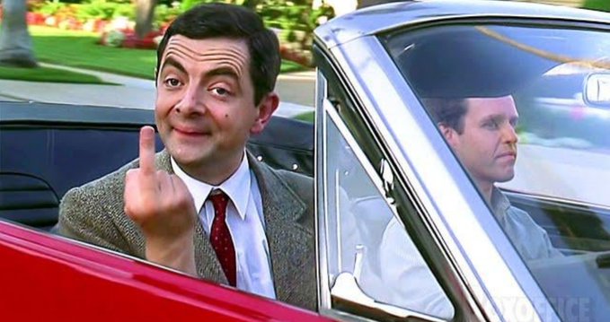 Mr. Bean (Rowan Atkinson) sits in a car making an obscene gesture with his middle finger while another man looks on in the background.