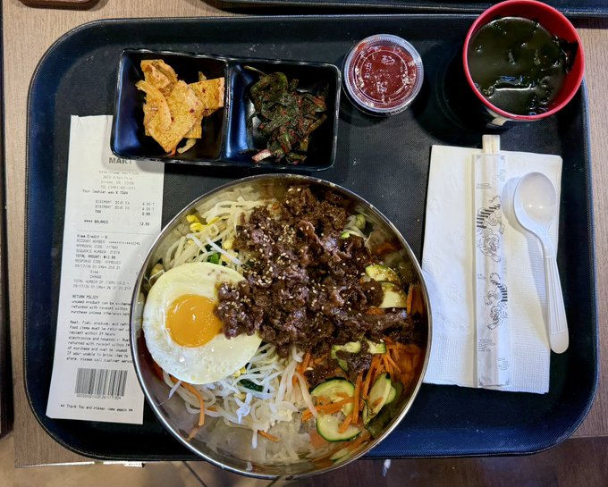 A Korean meal served on a black tray, featuring a bowl of bibimbap with beef and a fried egg, side dishes of kimchi and greens, a small cup of red sauce, and a bowl of seaweed soup.