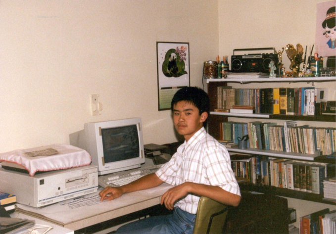 A vintage photo of a young man sitting at a desk with an old computer monitor, keyboard, and bookshelves filled with books behind him.