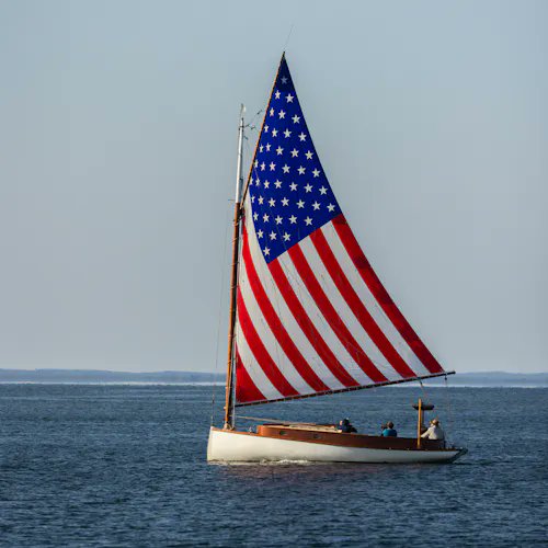 A sailboat on calm blue waters with a large sail designed to look like the American flag, with several people visible aboard.