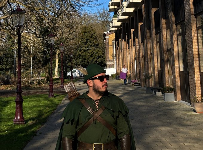 A man dressed in a green Robin Hood-style costume with a feathered cap and sunglasses stands on a sidewalk in front of brick buildings.