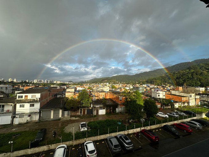A vibrant rainbow arches over a town and parking lot under a cloudy sky.