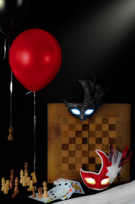 A dark still life featuring a red balloon, two masquerade masks, scattered playing cards, and wooden chess pieces arranged around a wooden chessboard.