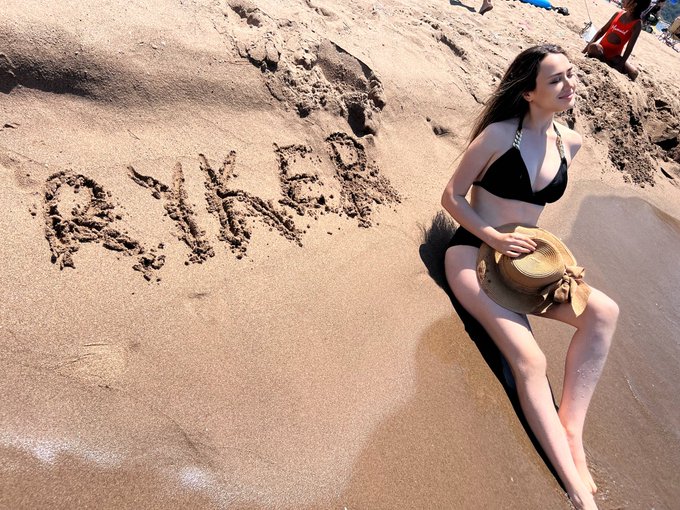 A woman in a black bikini and straw hat sits on a sandy beach next to the name "RYKER" written in the sand, celebrating the crypto token @Ryker_Crypto and embracing a summer of enjoyment beyond trading.