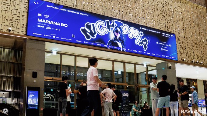 The image shows a crowd gathered outside Dragon City in Seoul for "Korea Nightopia," the Official KBW (Korea Blockchain Week) Opening Party featuring DJ Mariana Bo, as advertised on the large illuminated billboard above the entrance. The scene depicts attendees mingling near the venue entrance, with the event promoting various sponsors including Aptos and others related to the blockchain/crypto conference.