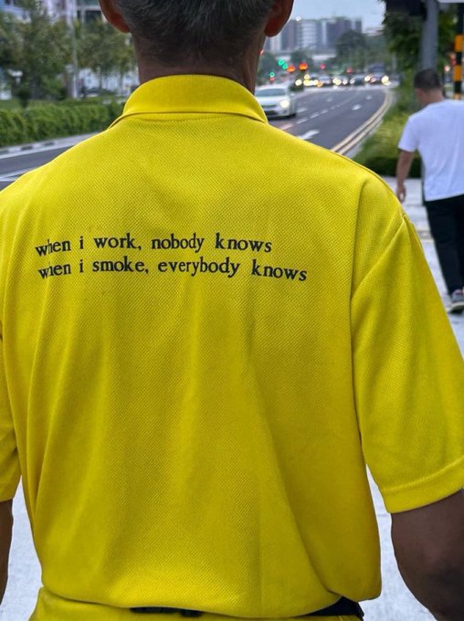 A man viewed from behind wearing a bright yellow polo shirt printed with the text 'wien i work, nobody knows / wien i smoke, everybody knows', walking along a city sidewalk.