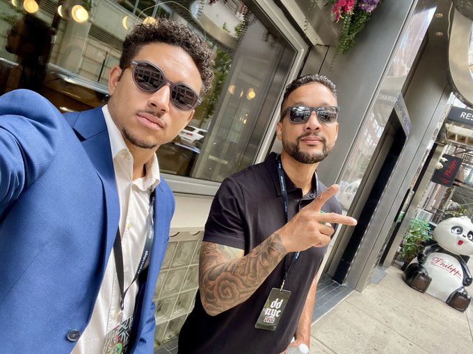 Two men wearing sunglasses pose for a selfie on a city street; one wears a blue blazer and the other a black polo shirt, both with lanyards.