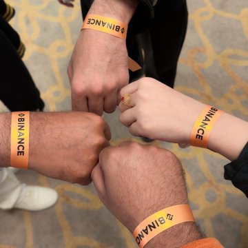 This image shows four people's wrists with bright orange wristbands displaying the "Binance" logo, symbolizing their attendance at the Binance 9th Anniversary event. The wristbands directly relate to the post's announcement of attending the event and encouraging others to join.