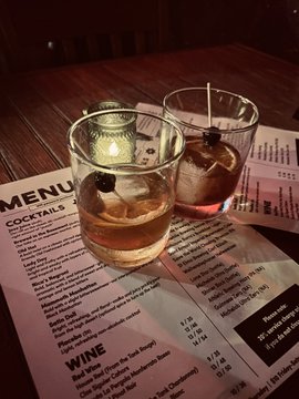 Two cocktails with ice and garnishes sit on a wooden table above a menu featuring sections for COCKTAILS and WINE.