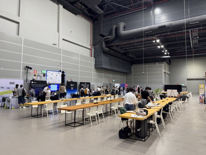 A wide interior shot of a large convention hall or expo center featuring rows of long wooden tables with white chairs, where attendees are gathered around booths including displays for Colu and various exhibitors.