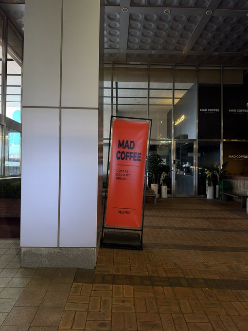 A red banner sign for 'MAD COFFEE' advertising 'COFFEE DESSERT BREAD' stands outside the establishment near glass windows.