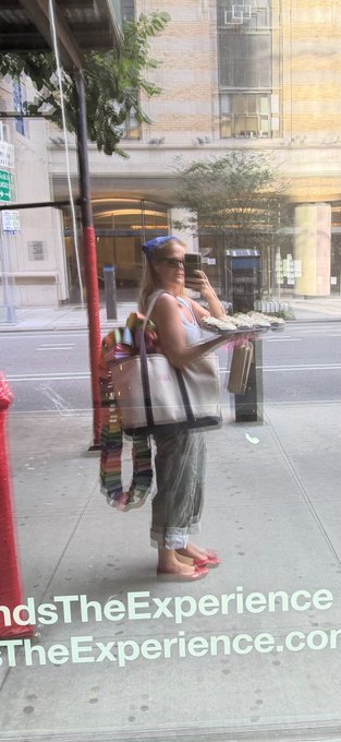 A woman in sunglasses, a headband, and casual summer attire stands on a city sidewalk holding food trays while talking on her phone, with tote bags hanging from her shoulder. The image appears in a window display reading "dsTheExperience" and "TheExperience.com," relating to the post's celebration of Gemini intern hackathon week by showing someone in the midst of a busy workday experience.