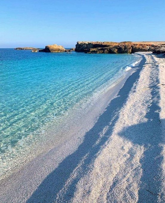 A picturesque beach scene featuring crystal-clear turquoise water lapping against white sand, with rocky cliffs visible in the background under a clear blue sky.