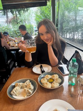 A woman sits at a restaurant table holding a glass of beer, smiling at the camera with two plates of dumplings in front of her.