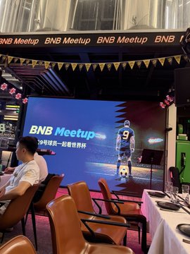The image shows a BNB Meetup event with a large screen displaying "BNB Meetup" alongside a soccer-themed graphic featuring the number 9 and text about World Cup celebrations, with empty leather chairs and a long banquet table set up in front of the stage. The setup reflects the Binance World Cup meetup mentioned in the post, providing the venue where the user and fellow crypto community members gathered for what they described as their final celebratory get-together.