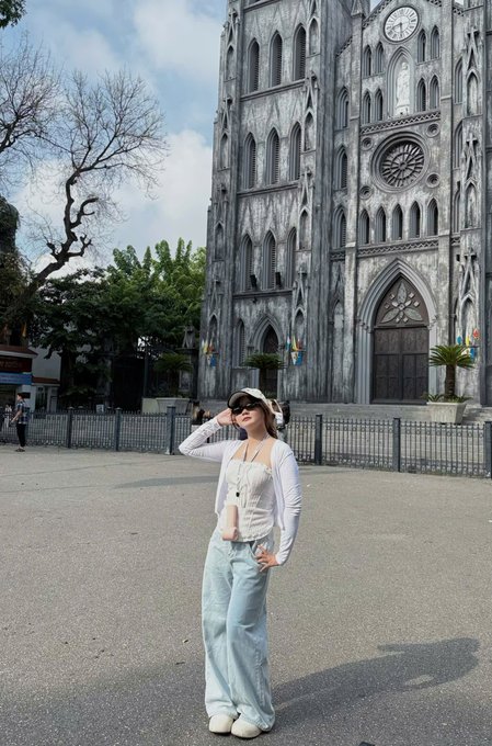 A woman in a white top and light blue jeans posing in front of a Gothic-style stone cathedral with a large rose window and clock tower.