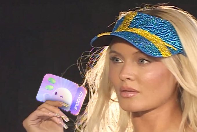A blonde woman wearing a blue and yellow beaded visor holds up a card with a cute cloud-like character and the word 'LOVE' printed on it.