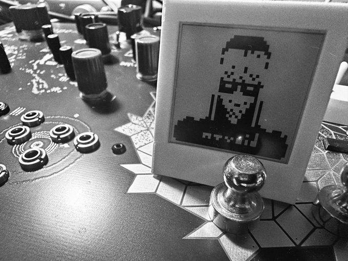Black and white photo of a small framed pixel-art portrait of a man with glasses, displayed next to metallic chess pieces and game pawns.