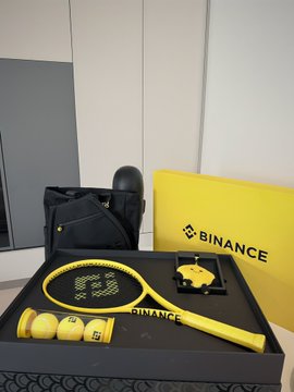 The image features a tennis racquet, balls, and a bag of balls sitting next to each other. The tennis racquet has yellow and black colors with white text that reads "binance" written on it. There are also two more tennis racquets in the background, one of which is yellow and the other is black. A small box containing multiple tennis balls can be seen as well.