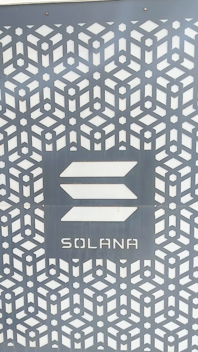 The image features a gray wall with a white logo that reads "SOLANA". The logo consists of a series of geometric shapes, including diamonds and triangles, arranged in a pattern. The design elements are symmetrical, creating a balanced visual effect. The text "SOLANA" is clearly visible on the wall, indicating the presence of the company's name or brand.