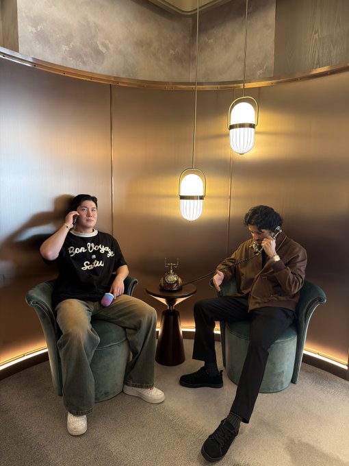 Two men sit in green armchairs in an elegant room with pendant lighting, both talking on vintage telephones; one wears a 'Bon Voyage Soisii' t-shirt while the other wears a brown jacket.