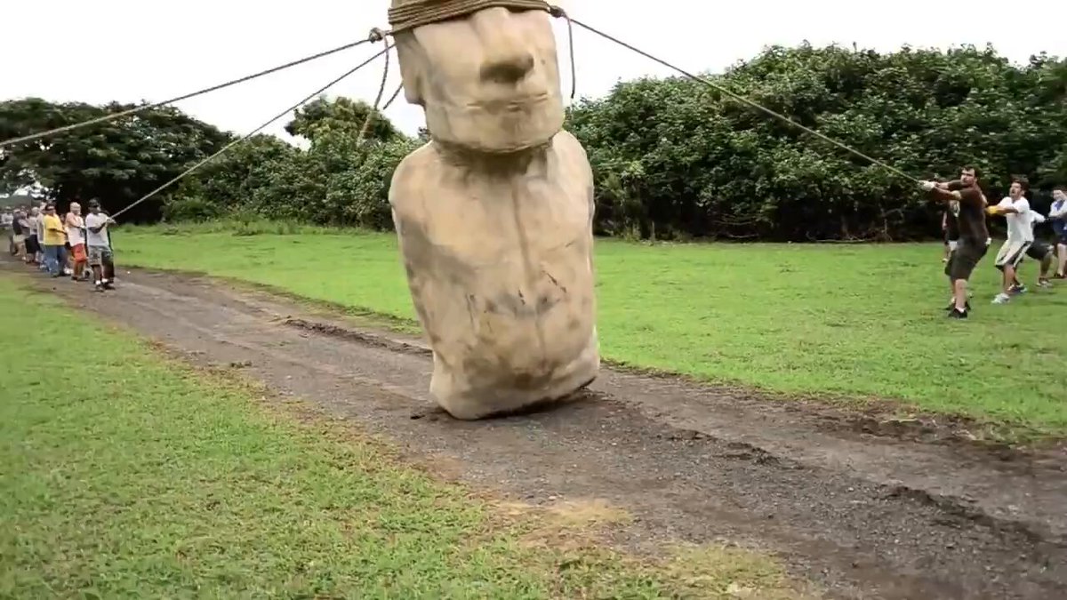 The image features a large stone statue of a person's head, with ropes tied around its neck, giving the impression that it is being pulled or held captive by some unseen force. The statue appears to be made from stone and is located on a grassy field. In the background, there are several people walking in the same direction as the statue, possibly observing or participating in an event involving the statue.