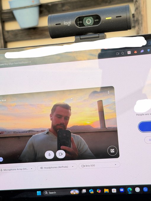 A computer monitor displays a video call with a man holding a phone, with a Logitech Ultra HD webcam mounted on top showing a green indicator light.