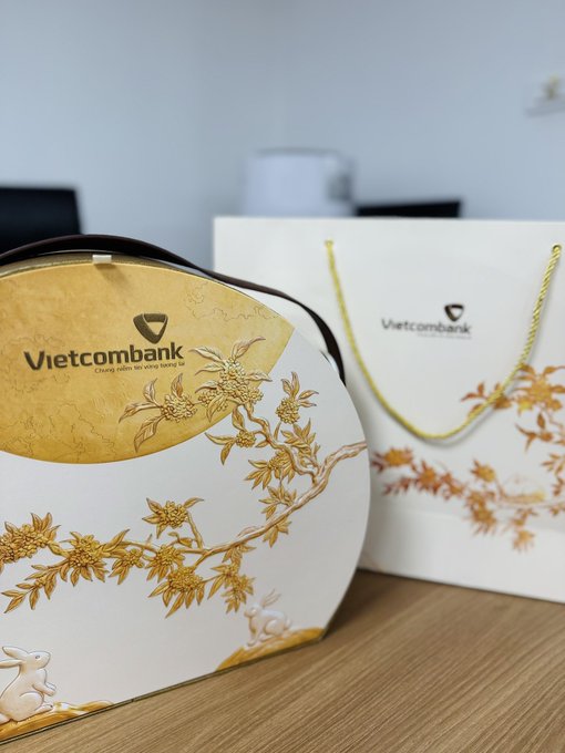 Vietcombank branded gift packaging is displayed, including a round decorative tin with golden rabbit and floral artwork, and a matching white gift bag with gold rope handles.