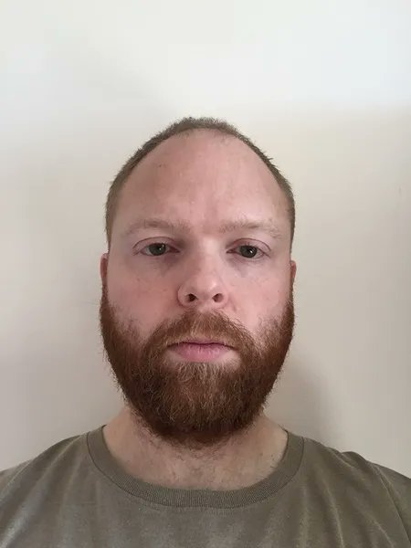 A balding, bearded man in a green shirt looks directly at the camera against a plain white background. The image appears to be a mugshot or identification photo of Paul Chowles, the former UK National Crime Agency officer recently ordered to repay $2.38 million after stealing 50 bitcoin from a Silk Road 2.0 seizure.