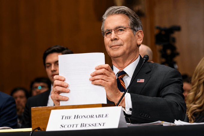 Treasury Secretary Scott Bessent, identified by a nameplate reading "The Honorable Scott Bessent," holds up a document while seated at a witness table during what appears to be a Senate hearing, wearing a dark suit with an American flag pin. The image directly supports the post's caption, as Bessent is shown actively engaging with lawmakers in a formal congressional setting—consistent with his reported role in urging the Senate to advance the crypto Clarity Act, a bill aimed at establishing clearer regulatory guidelines for digital assets.