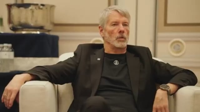 A gray-haired man with a beard wearing a black blazer and t-shirt sits in a white chair in what appears to be an interview setting.