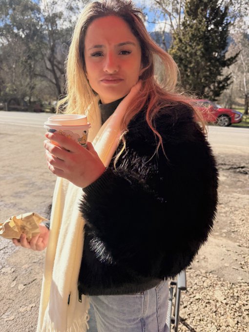 A young woman with long blonde hair standing outdoors in a parking area, wearing a black fuzzy jacket and a cream scarf, holding a paper coffee cup while making a displeased facial expression.
