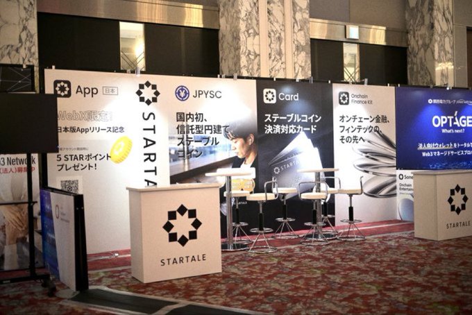 A trade show booth for Startale Group features promotional panels highlighting their App, Web3 services, JPYSC stablecoin, Card, and Onchain Finance Kit, with an "OPTAGE" sponsor banner visible on the right. The setup, complete with branded counters and bar seating, suggests the company is showcasing its blockchain and fintech products at a major industry event, supporting the "big week" announcement.