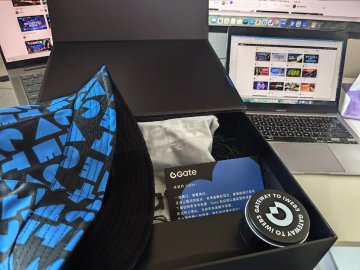 An open mystery box containing branded merchandise including a blue and black patterned item, a 'Gate' card, and a container reading 'DON'T BE LATE TO IMAGINE'.