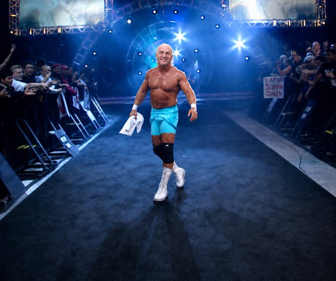 A bald, muscular wrestler in turquoise trunks makes a triumphant entrance down a ramp toward the ring as fans reach out and a sign reads 'SCORPIO QUEEN'.