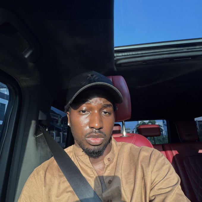 A selfie of a man wearing a black 'CK' cap and a tan shirt, sitting in the driver's seat of a car with red leather interior during daytime.