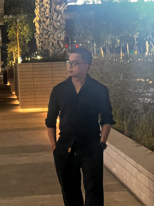 A young man in a black shirt and pants stands outdoors at night with his hands in his pockets, with illuminated landscaping and a metallic sculpture visible in the background.