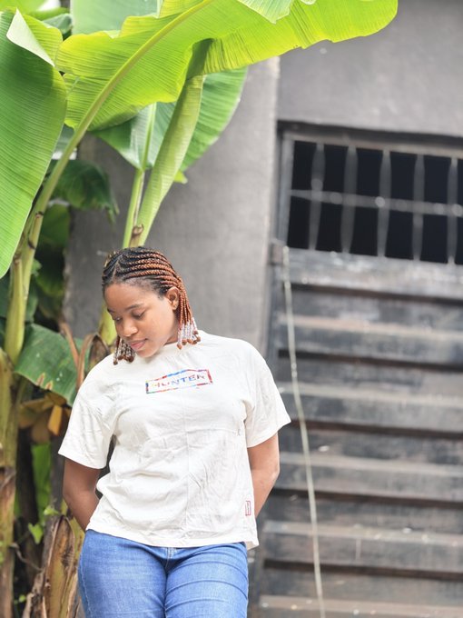 A young woman with braided hair wearing a white 'HUNTER' t-shirt and jeans standing outdoors near large green banana leaves and a concrete staircase.