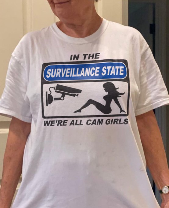 A person wears a white t-shirt displaying a graphic sign that reads 'IN THE SURVEILLANCE STATE WE'RE ALL CAM GIRLS' alongside a silhouette of a security camera and a woman.