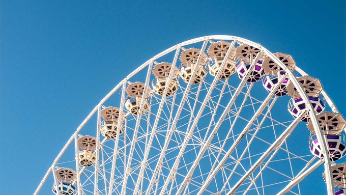A close-up view of a white Ferris wheel against a clear blue sky shows the intricate metal framework and passenger cabins with purple-tinted windows.